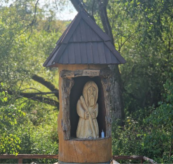 Tradycyjna, drewniana kapliczka przydrożna z rzeźbą, w otoczeniu zieleni.