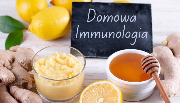 domowe składniki na odporność: świeży imbir w kawałkach i starty w miseczce, miseczkę miodu, połówkę i całe cytryny. W tle znajduje się mała czarna tabliczka z napisem "Domowa Immunologia".