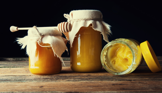 Trzy słoiki miodu na drewnianym stole – dwa z dekoracyjnymi kapturkami i miodarką, jeden otwarty ze skrystalizowanym miodem.