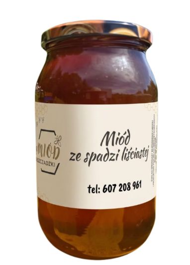 Słoik ciemnego miodu ze spadzi liściastej na tle rozmytej scenerii ogrodowej. Na etykiecie widnieje napis 'Miód ze spadzi liściastej', logo pasieki i numer telefonu: 607 208 961.